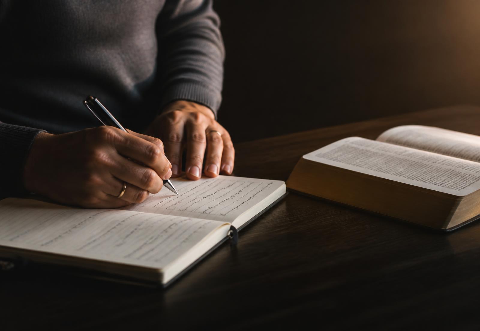 Mãos escrevendo em um caderno ao lado de um livro aberto sobre mesa de madeira
